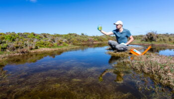 A person holds a jar while sitting near a pond